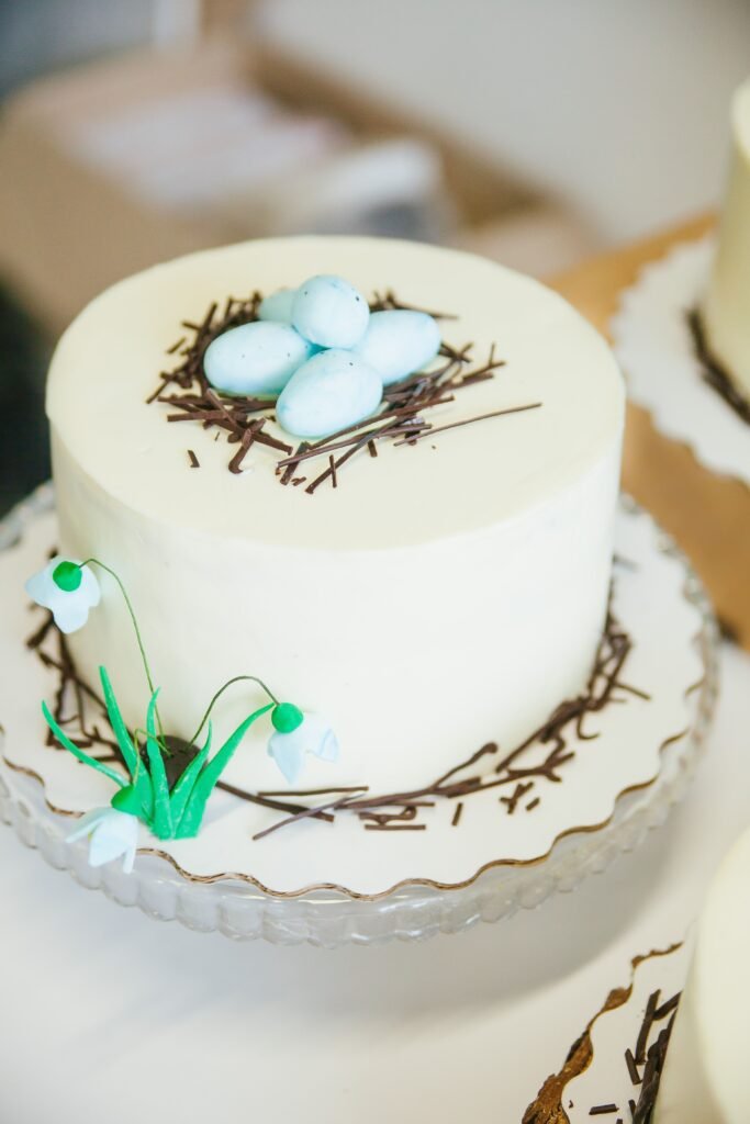 Elegant white Easter cake with blue candy eggs, chocolate nest decoration, and spring flower details.