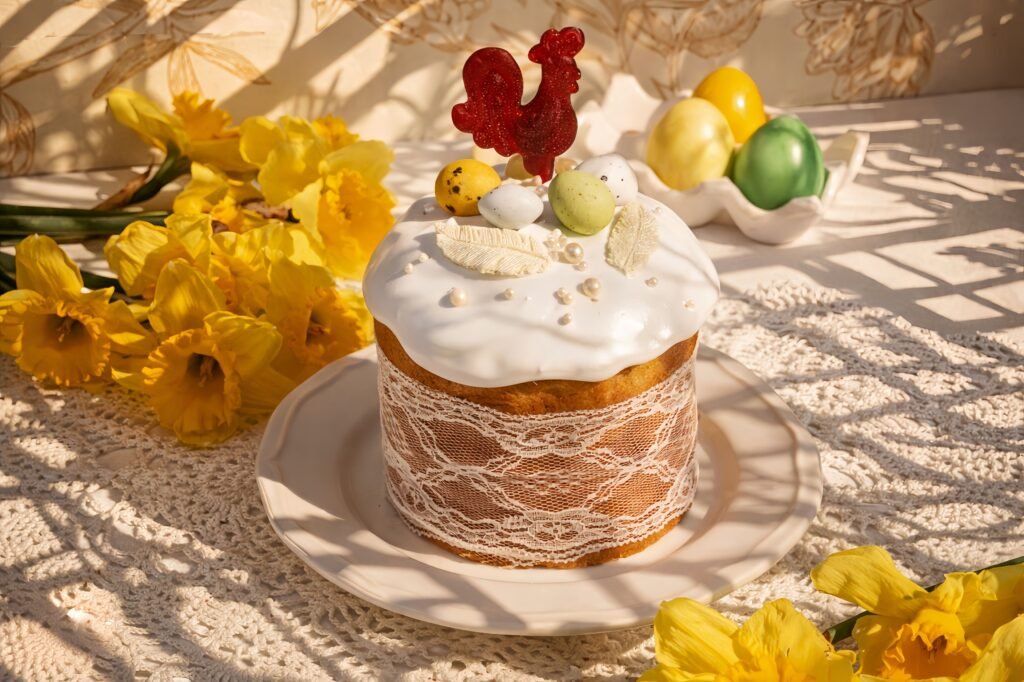 Rustic Easter cake with white glaze, lace wrap, colorful candy eggs, rooster topper, and yellow spring flowers.